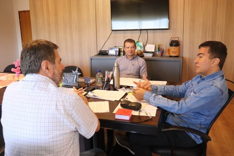 Three men sit around a desk in a wood-paneled office, discussing with scattered papers, notebooks, and pens on the table, backs and side profiles visible.