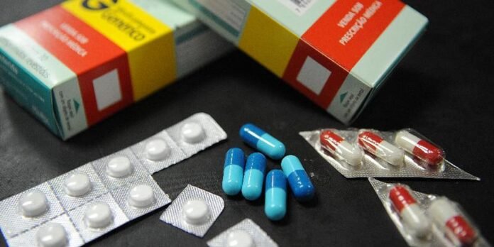 Assorted medications on a dark surface: blue and white capsules, white tablets in blister packs, and colorful pill boxes in the background.