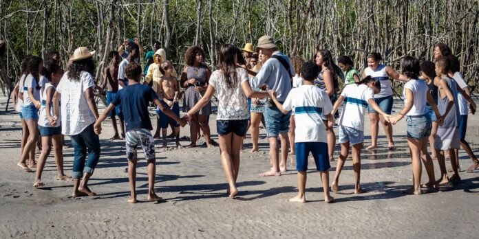 Group of kids and adults holding hands in a circle on a sandy beach with mangrove trees in the background.