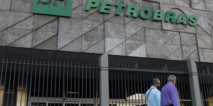Exterior of a Petrobras building with a green sign and geometric concrete wall, a tall metal fence, and two men walking by.