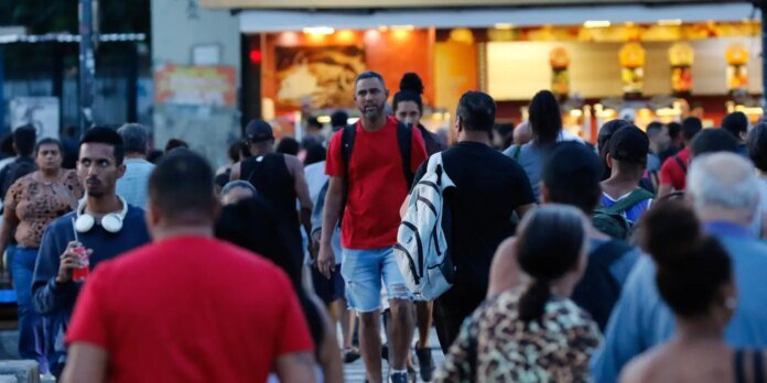 Crowded city street at dusk with a food stand in the background and a man in a red shirt and backpack walking toward the camera.