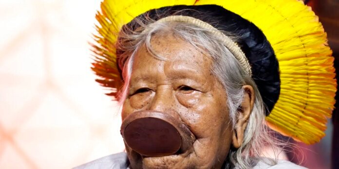 Close-up of an elderly person wearing a bright yellow feathered ceremonial headpiece and a large brown circular nasal adornment.