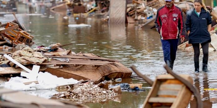 Two people in rain jackets walk through waist-deep floodwater past piles of debris and damaged buildings.