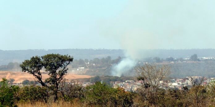 Smoke rising over a town in a dry, grassy landscape; trees in the foreground with a distant hill range behind.