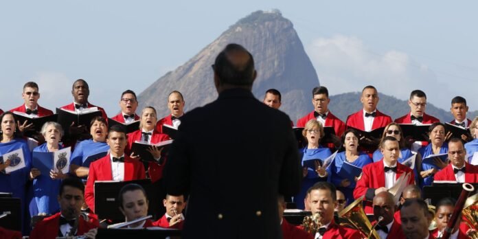 Choir performing outdoors, viewed from behind the conductor in a dark suit; singers wear red jackets and blue dresses, mountain in the background