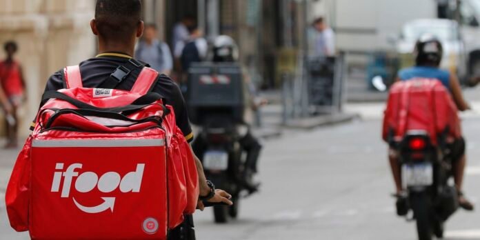 Two food delivery riders on motorcycles with red insulated bags labeled iFood in a city street, seen from behind.