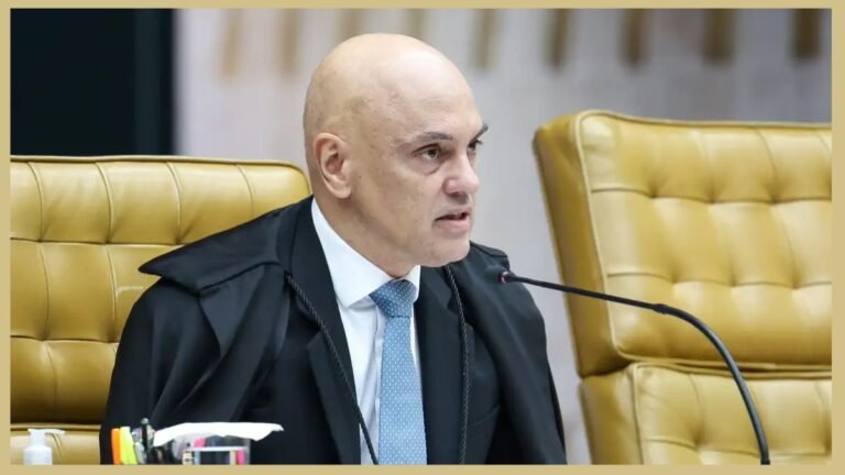 Bald official in a black robe and blue tie speaks into a microphone at a formal hearing table.