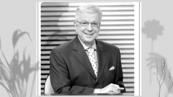 Black-and-white portrait of a smiling older man in a dark suit with a polka-dot tie, seated with hands folded in front.