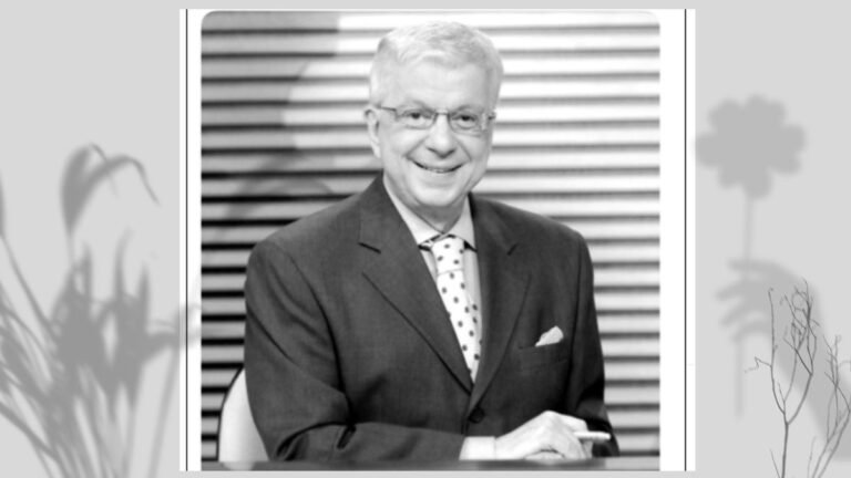 Black-and-white portrait of a smiling older man in a dark suit with a polka-dot tie, seated with hands folded in front.