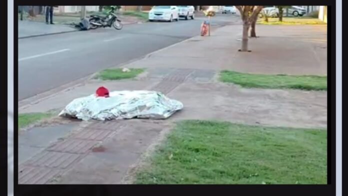 A body or object covered with a light blanket on a sidewalk beside a street, with a red item on top.