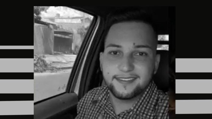 Black-and-white photo of a smiling man with short hair and a beard, wearing a checkered shirt, sitting in a car.
