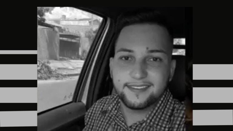 Black-and-white photo of a smiling man with short hair and a beard, wearing a checkered shirt, sitting in a car.