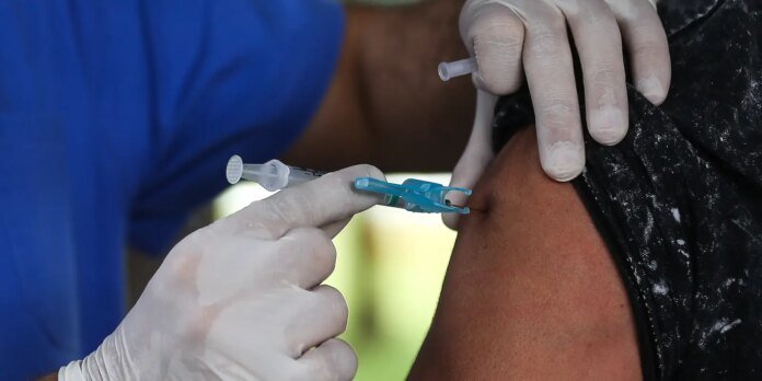 Healthcare worker wearing gloves administers an injection into a patient’s upper arm; syringe visible.