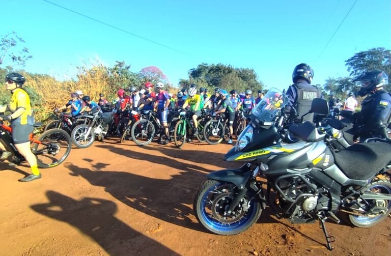 A large group of riders wearing helmets and colorful cycling outfits gathered on a dirt road, with a police motorcycle in the foreground on the right.
