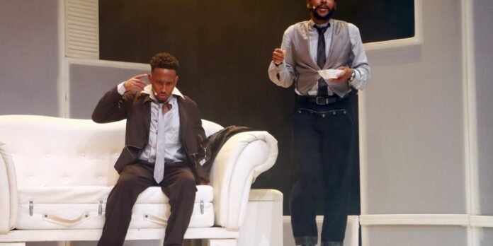 Theatrical scene with two actors: a man in a suit sitting on a white couch making a dramatic gesture, while another man in a shirt and tie stands nearby holding a bowl and spoon as if performing.