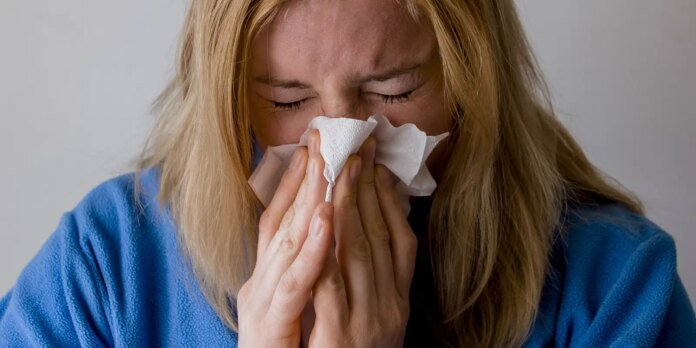 Woman with blonde hair wearing a blue robe blowing her nose into a tissue with eyes closed.