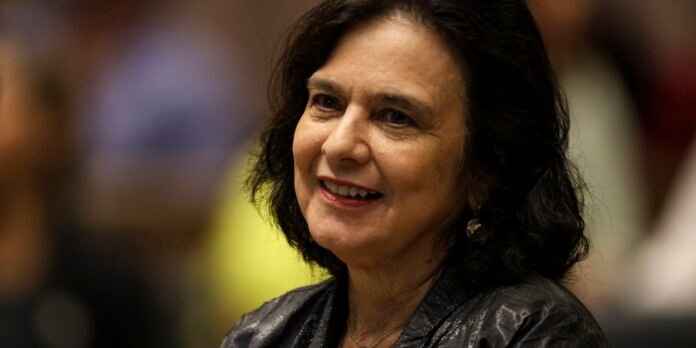Smiling woman with dark hair in a metallic jacket at an indoor event, facing the camera.