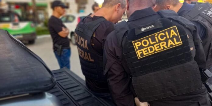 Two federal police officers in black tactical vests marked POLICIA FEDERAL stand beside a vehicle on a street, conversing or surveying a scene in the background.
