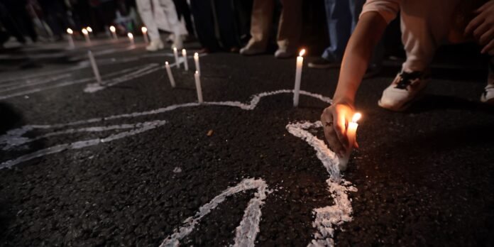 People at a nighttime vigil placing lit candles on the street, creating a chalk-like outline on the pavement.