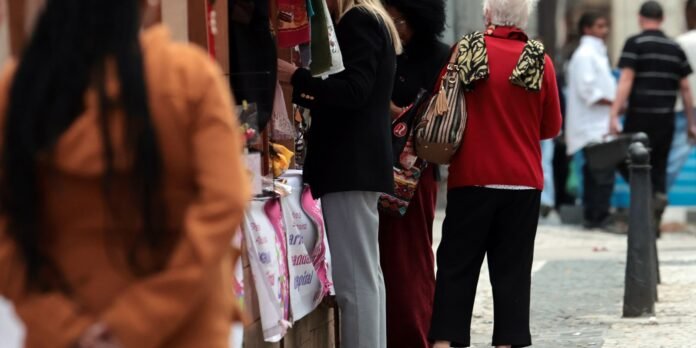 Shoppers browse an outdoor market stall with colorful bags, fabrics, and accessories along a city sidewalk.