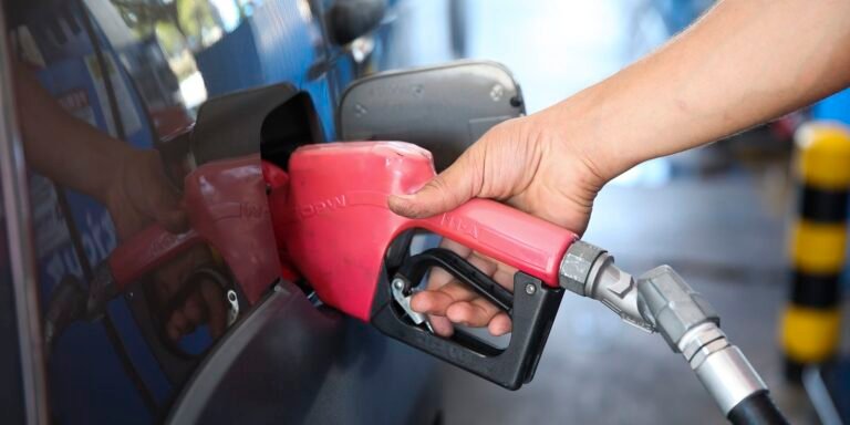 Person holding a red gas pump nozzle and inserting it into a car’s fuel tank.