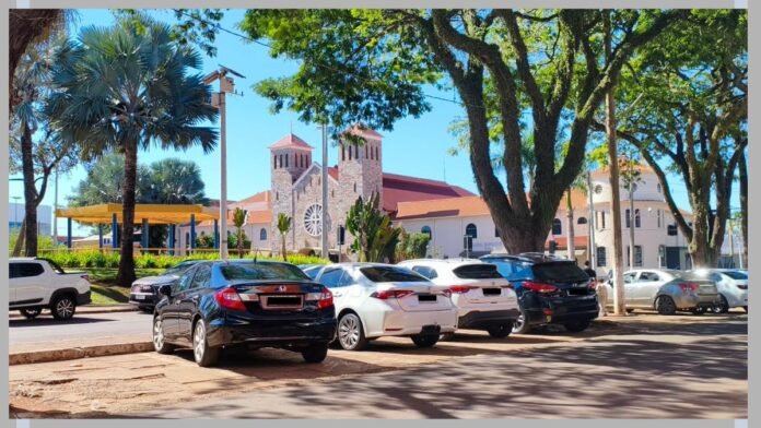 Line of parked cars along a tree-lined street with a stone church in the background under a bright blue sky