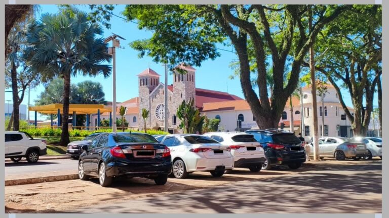 Line of parked cars along a tree-lined street with a stone church in the background under a bright blue sky