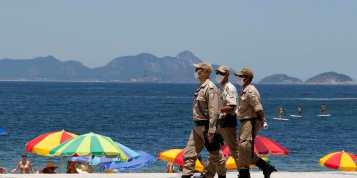 Three police officers in tan uniforms and face masks patrol a sunny beach with colorful umbrellas and sunbathers in the foreground.