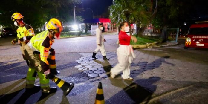Nighttime scene with firefighters in high-visibility gear handling equipment on a lit street, while a woman in red and a person in white protective suit walk by.