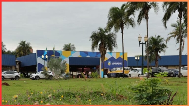City hall (Prefeitura de Dourados) with a blue–yellow geometric mural, cars parked outside and palm trees in a grassy area.