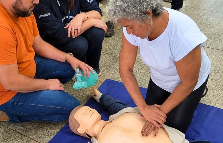 CPR training scene: a woman performs chest compressions on a life-size manikin while another person holds a bag‑valve mask nearby.