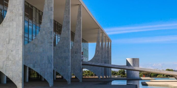 Modern marble building with tall angled columns, a bridge-like walkway, and a blue sky reflecting in a pool below.