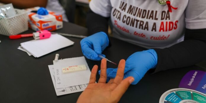 Healthcare worker in blue gloves performs a finger-prick blood test on a person's fingertip at an AIDS awareness event.