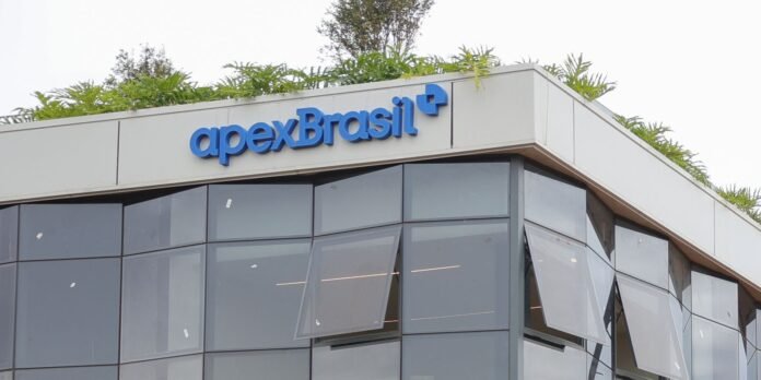Modern office building with dark glass panels and a blue 'apexBrasil' sign on the top edge, with greenery on the roof.