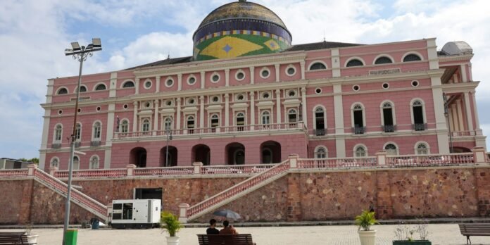 Pink neoclassical building with a colorful mosaic dome atop a stone retaining wall, set in a sunny plaza; two people sit on a bench in the foreground.