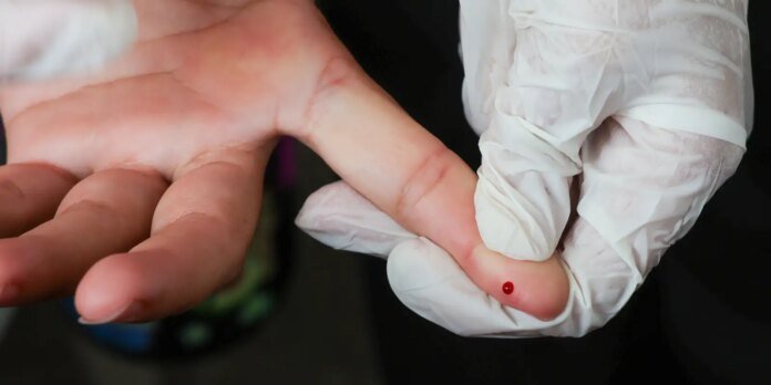 Close-up of a gloved hand pricking a fingertip with a tiny drop of blood visible on the skin's surface.