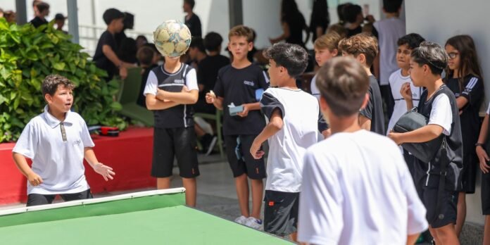 Group of students watches as a boy in a white polo prepares to volley a soccer ball onto a green table during a school event.