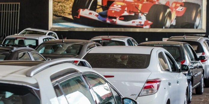 Indoor car showroom/garage with rows of parked cars, white sedan in foreground and a large wall mural of a red race car in the background.