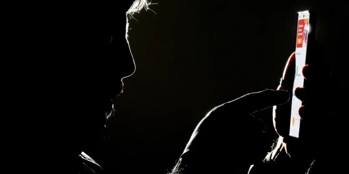 Silhouette of a person in a dark room holding a bright smartphone toward their face.