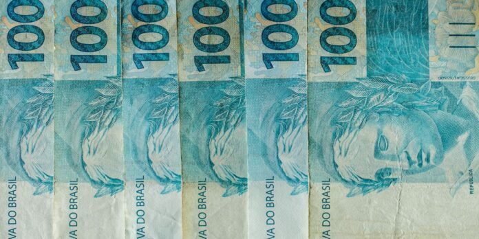 Blue 100 reais banknotes lined up side by side, showing the large '100' numerals and a portrait on the right with leafy patterns visible across the notes.