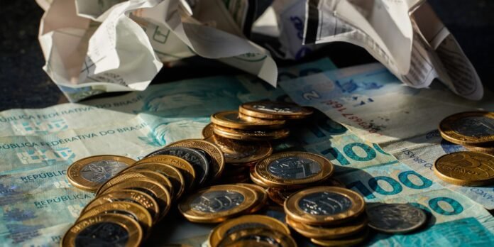 Coins and Brazilian reais scattered on banknotes with crumpled papers above, suggesting cash handling or money on a surface.