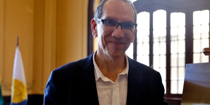 Smiling man in a dark suit and glasses indoors, with a flag in the background.