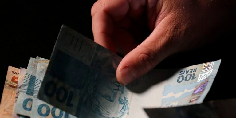 Close-up of a hand fanning Brazilian real banknotes on a dark surface.