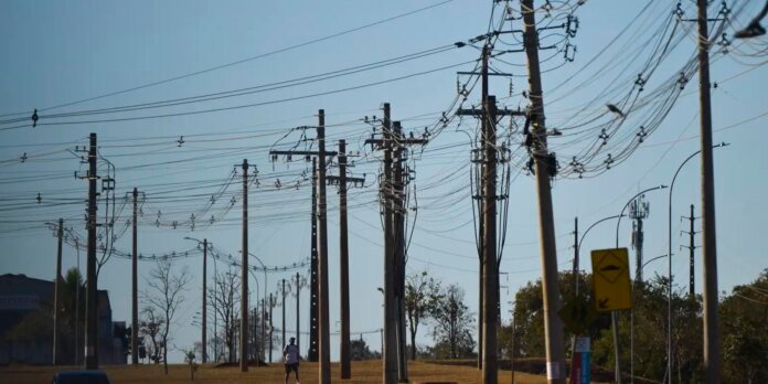 Row of tall wooden utility poles with many overhead wires along a street; a lone pedestrian is visible in the distance under a clear blue sky.