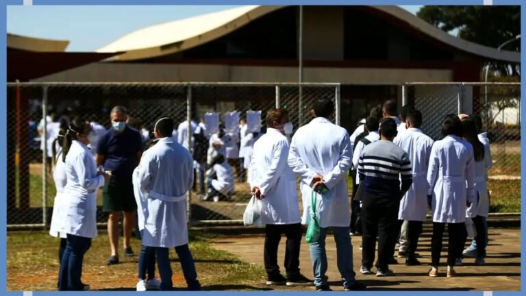 Group of people in white lab coats standing in line outside a fenced building; several wear masks amid a sunny outdoor setting.