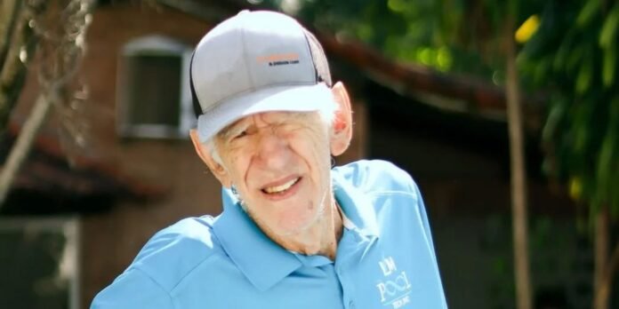 Older man wearing a light blue polo with a logo and a gray cap, smiling in a sunny outdoor setting.