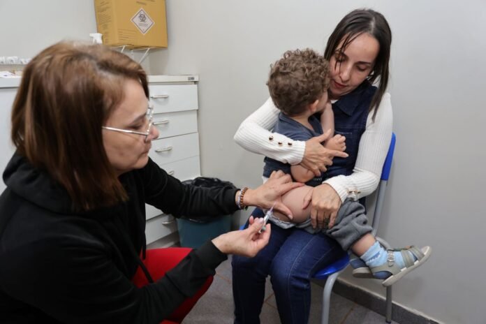 Healthcare professional giving a vaccination to a toddler seated on a parent's lap in a clinic room.