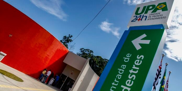 Exterior view of a UPa 24h Unidade de Pronto Atendimento sign with a green panel and a left arrow, a red curved wall, and people queuing at the entrance under a blue sky.