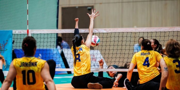 Volleyball match: a player in a yellow jersey (number 3) reaches up to block the ball at the net while others watch, one player on the floor nearby.
