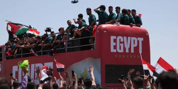 A crowded red tour bus labeled 'EGYPT' with soccer players waving to cheering fans and waving Egyptian flags from the top deck.